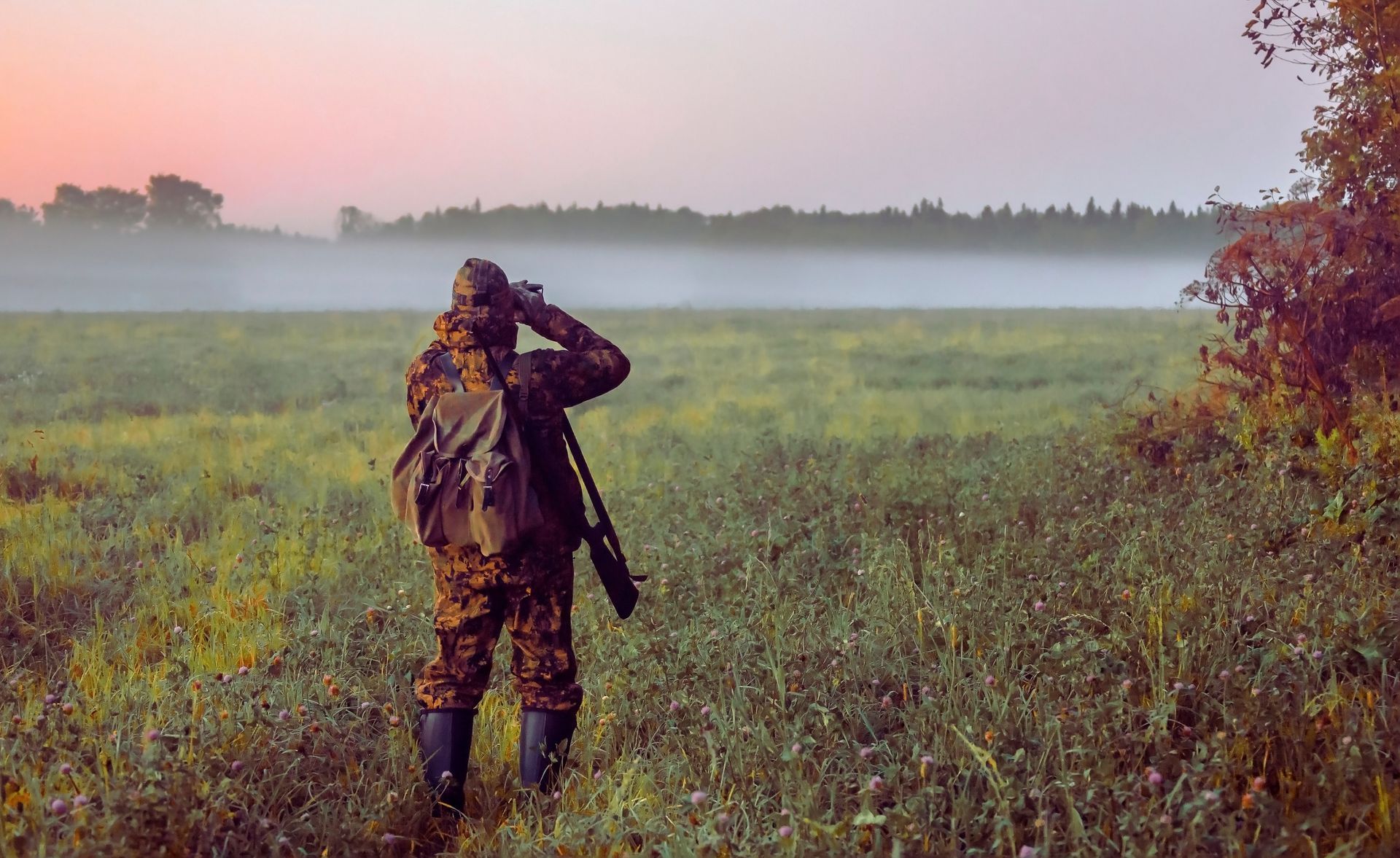 Hunter in camouflage gear, looking through binoculars in a misty field at sunrise.