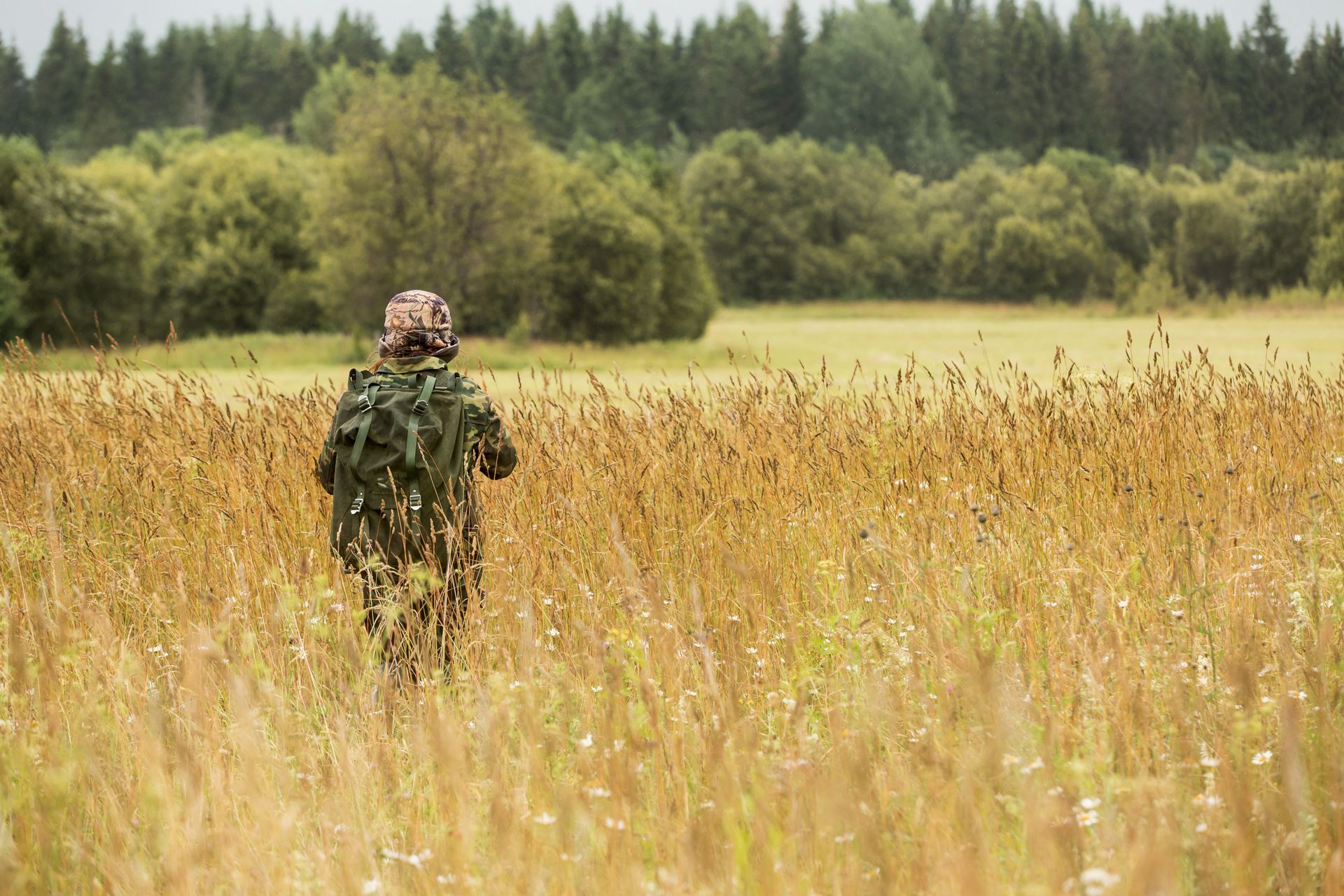 Person in camouflage gear walking through a field of tall grass toward a treeline.
