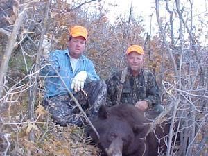 Two men in hunting attire pose with a large, dark bear in a wooded area.