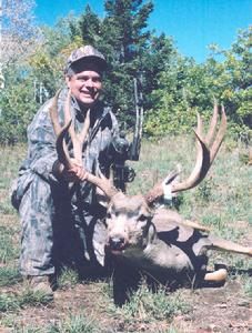 Hunter kneels beside a large mule deer buck in a wooded area, smiling and holding the deer's antlers.