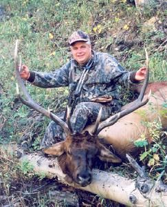 Hunter in camouflage sits on a fallen elk, holding up antlers in a wooded area.