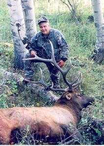 Man in camouflage kneels beside a large elk with large antlers in a wooded area.
