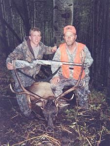 Two people pose with a harvested elk in a forest. One wears camouflage, and the other wears an orange vest. They hold rifles.