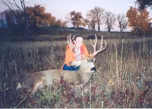 Woman in orange jacket seated on a large buck deer in a grassy field.