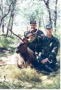 Two men pose with a dead elk in a wooded area. They wear hats and camouflage.