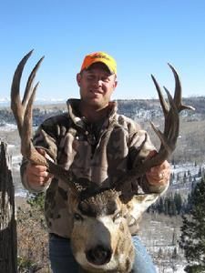 Man in camouflage holding large deer antlers, with snowy mountain backdrop.