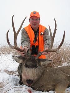 Hunter in orange vest holding large deer antlers, cloudy field in background.