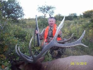 Hunter kneeling beside a large elk, holding rifle. Wearing blaze orange vest in a grassy field.