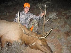 Hunter in camo kneels next to a dead elk, holding its antlers in a dark outdoor setting.