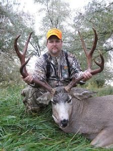 Hunter kneeling with a large buck. Hunter wears camouflage and an orange hat, holding the buck's large antlers in a grassy outdoor setting.