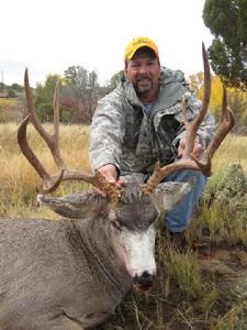 Man in camouflage jacket kneeling beside a large mule deer with large antlers. Outdoor setting.