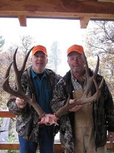Two men in hunting gear hold a large deer antler rack outdoors, smiling.