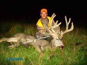 Man kneels next to a dead buck with large antlers, rifle across it. Green grass, night.