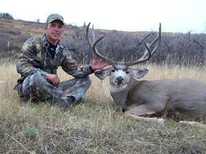 Hunter kneels beside a large mule deer buck in a field, smiling. Deer has large antlers.