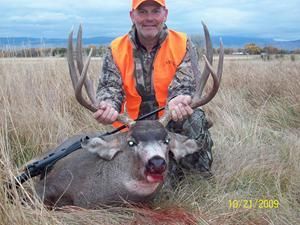 Hunter in orange vest kneels next to a dead deer with large antlers, rifle nearby; outdoor setting.