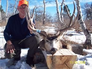 Hunter kneels beside a mounted deer head with large antlers. Orange hat, blue shirt, snowy outdoors.