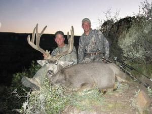 Two people pose with a large buck deer in outdoor setting with hunting gear.