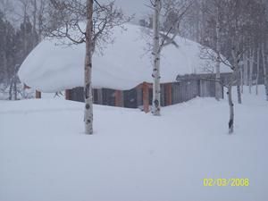 Snow-covered wooden building in a snowy landscape, trees in the foreground.