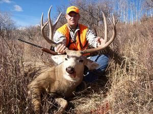 Hunter in orange vest kneels beside a large buck with antlers in a grassy field.