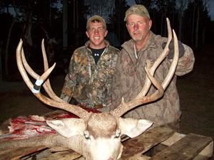 Two men pose with a large deer they hunted. The deer is on a wooden surface with a light-colored coat and large antlers.