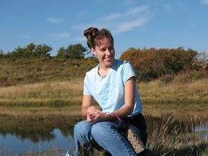 Woman in blue shirt and jeans sits by water, smiling in a natural setting.