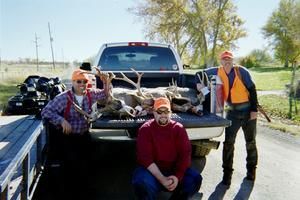 Three hunters pose with deer in a truck bed. Orange vests, hats. Sunny outdoor setting.