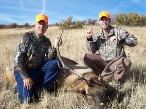 Two hunters kneeling with elk on field, both wearing orange hats, one pointing up, other making 
