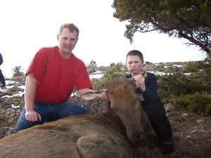 Two people with an elk they appear to have hunted. Man in red shirt, boy, snowy hillside.