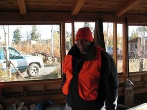 Man in orange hunting vest and hat inside a wooden cabin, truck in background.