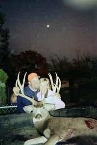 Couple poses with a large buck they killed. Nighttime shot. Man in orange hat, woman smiles. Deer has large antlers.