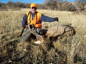 Hunter kneels by a downed elk in a field. He wears orange vest and hat. Rifle is visible.