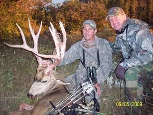 Two men in camouflage pose with a large buck and bow.