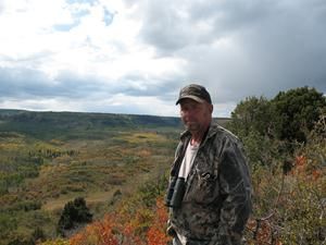 Man in camouflage, binoculars, standing in a hilly landscape with autumn foliage and cloudy sky.