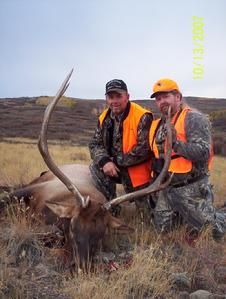 Two men in hunting gear kneel near a dead elk with large antlers in a field.