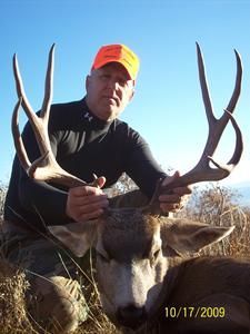 Hunter kneeling with a large buck deer, holding its antlers; blue sky background.