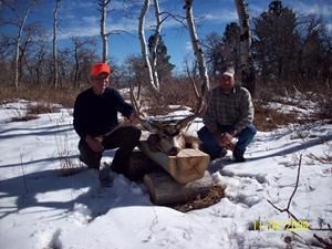 Two men squat beside a deer head trophy on logs in snowy woods. One man wears orange hat.