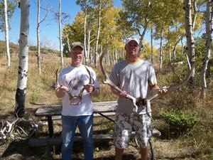 Two men outdoors holding large antlers. Trees in background, sunny day.