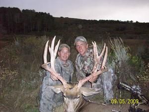 Two people in camo pose with a large buck deer, possibly after a hunt, in a natural setting.
