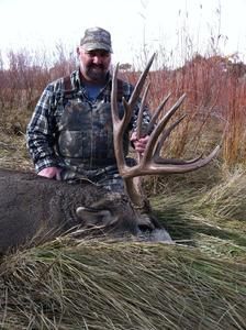 Man in camo with a large buck. The buck has a large rack of antlers and is lying in tall grass.