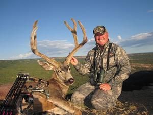 Hunter kneels beside a large buck with impressive antlers, holding binoculars, outdoors.
