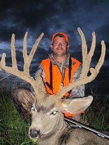 Hunter in orange vest kneels, holding large buck with massive antlers, night setting.