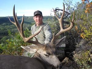 Man with a deer he hunted, holding its large antlers. He is wearing camo in a forest setting with a bow.