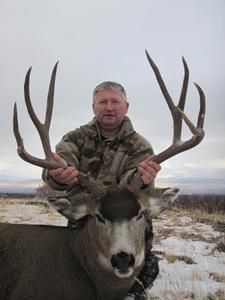 Man in camouflage holds up a large mule deer buck with impressive antlers outdoors.
