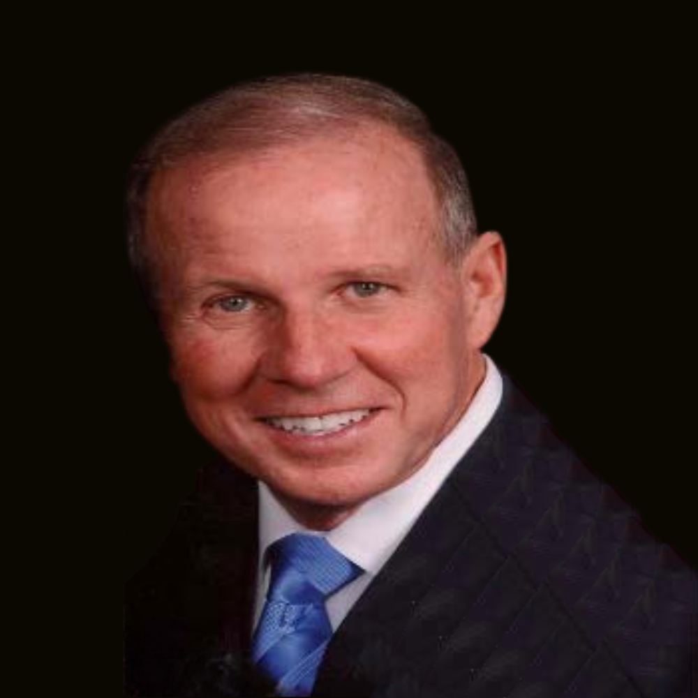 A man smiling in a dark suit, white collared shirt, and blue tie against a black background.