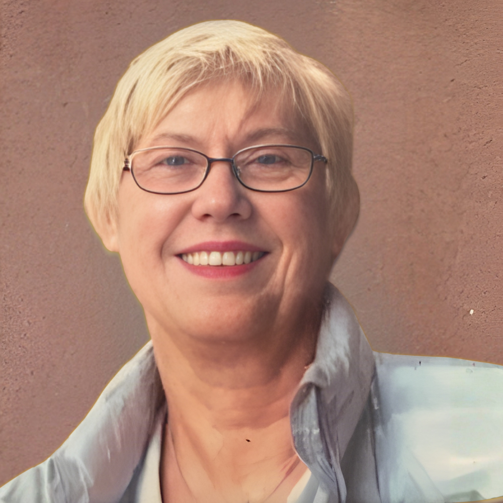 A person with short blonde hair and glasses smiles warmly against a neutral background, wearing a light blue jacket.