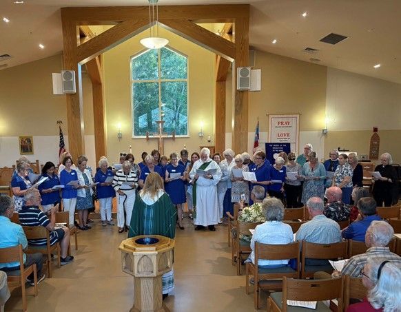 A group of people standing and singing in a church sanctuary, with a clergy member leading from the center.