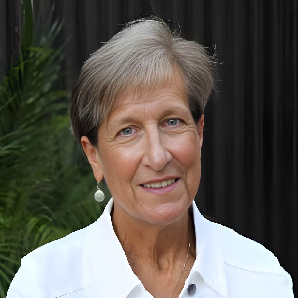 Headshot of a person with short, light-brown hair and silver earrings, wearing a white shirt against a dark background.