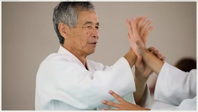 Een man in een witte judogi demonstreert een vechtsporttechniek, binnenshuis.