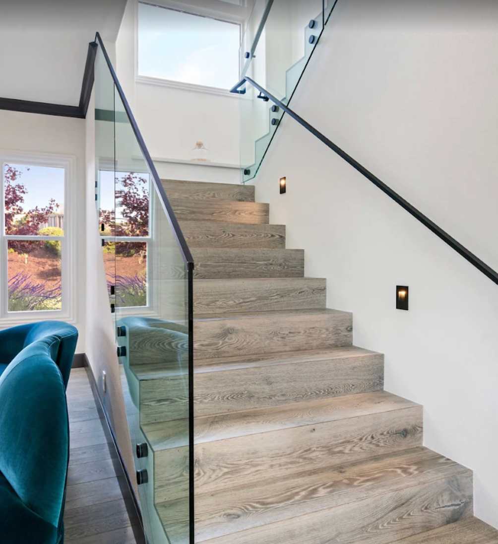 Wooden staircase with glass railing. Wall-mounted lighting, black handrail. Windows with outdoor view.