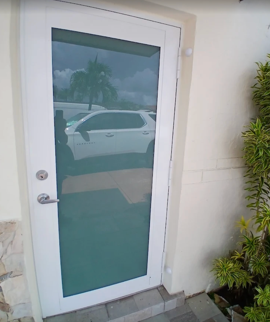 White framed glass door; cars and palm tree reflection. Light beige walls; greenery on right.